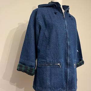 May include: A blue denim jacket with a hood and a zipper closure. The jacket has three-quarter sleeves and a green and black plaid lining on the cuffs and hood. The jacket is displayed on a mannequin.