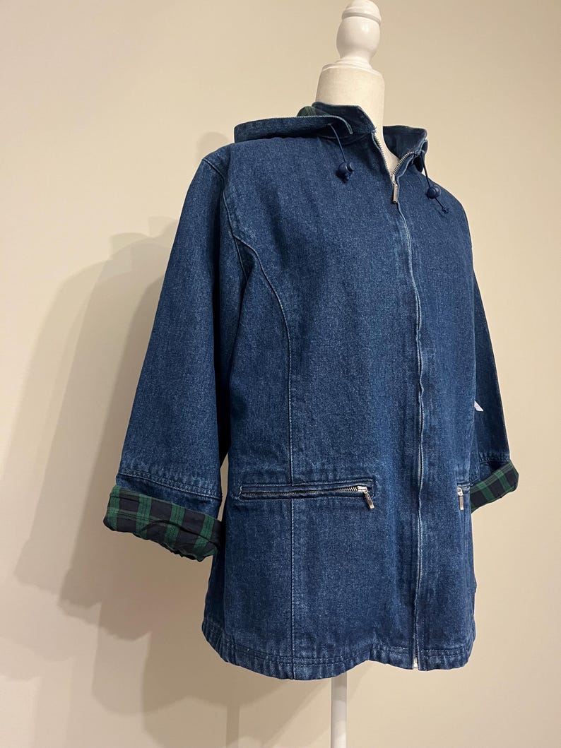 May include: A blue denim jacket with a hood and a zipper closure. The jacket has three-quarter sleeves and a green and black plaid lining on the cuffs and hood. The jacket is displayed on a mannequin.