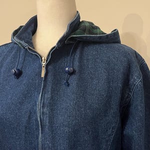 May include: A blue denim jacket with a hood. The hood lining features a green and black plaid pattern. The jacket has a zipper closure and blue drawstrings with blue toggles. The jacket is displayed on a mannequin.