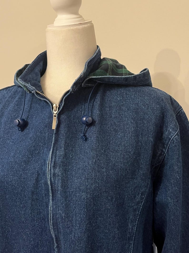 May include: A blue denim jacket with a hood. The hood lining features a green and black plaid pattern. The jacket has a zipper closure and blue drawstrings with blue toggles. The jacket is displayed on a mannequin.
