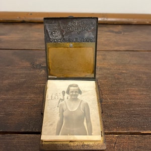 May include: An antique metal photo holder, open to display a black and white photograph of a woman in a swimsuit at the beach. The holder has a hinged top with embossed text and a brass-colored interior.