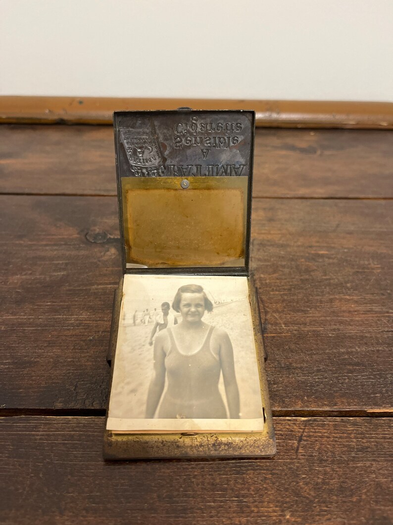 May include: An antique metal photo holder, open to display a black and white photograph of a woman in a swimsuit at the beach. The holder has a hinged top with embossed text and a brass-colored interior.