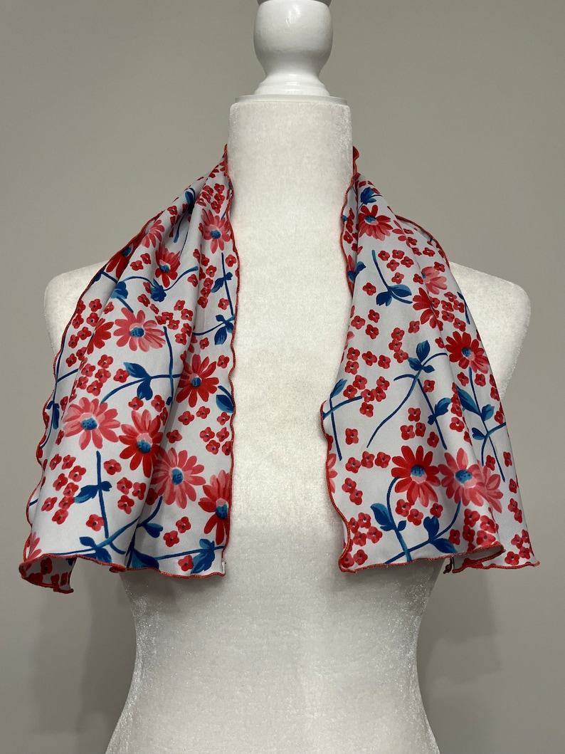 May include: A white scarf with a red and blue floral pattern. The scarf has a scalloped edge and is displayed on a white mannequin.