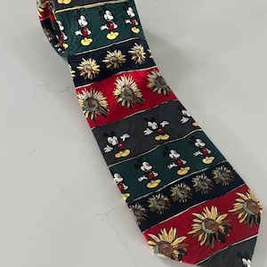 Puede incluir: Una corbata estampada con rayas alternas en gris oscuro, verde, rojo y negro. La corbata está decorada con imágenes de Mickey Mouse y girasoles. La corbata está hecha de un material brillante.