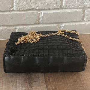 May include: A black rectangular handbag featuring a quilted grid pattern across the front surface. The bag includes a long, thick gold-tone metal rope-style chain strap that rests on top of the bag. The base and sides are smooth black material.
