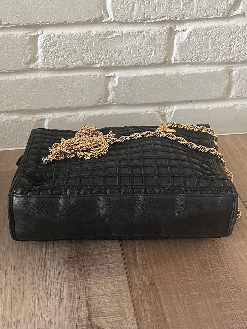 May include: A black rectangular handbag featuring a quilted grid pattern across the front surface. The bag includes a long, thick gold-tone metal rope-style chain strap that rests on top of the bag. The base and sides are smooth black material.