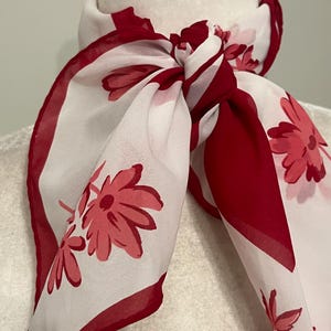 May include: A white and red floral scarf tied in a knot. The scarf features a repeating pattern of red flowers on a white background, with a solid red border. The scarf is draped around a white mannequin head.