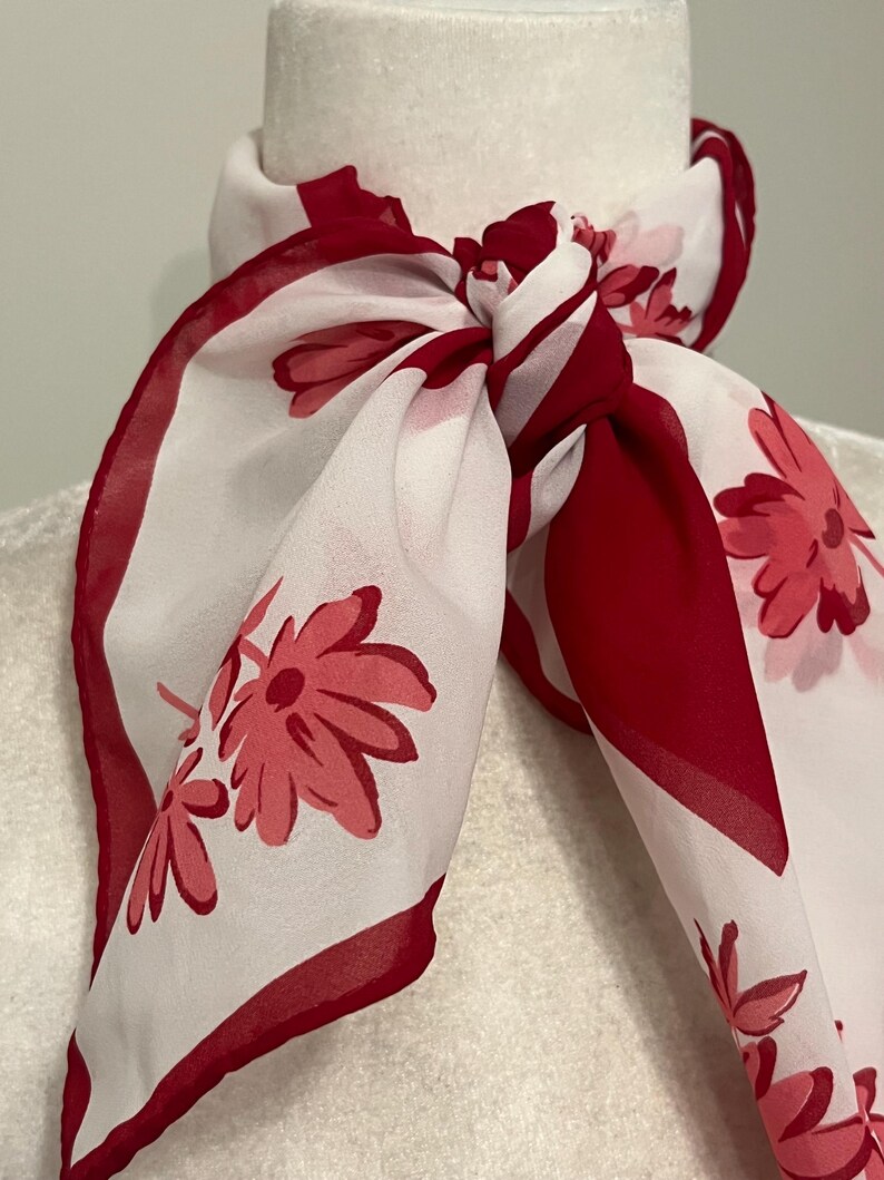 May include: A white and red floral scarf tied in a knot. The scarf features a repeating pattern of red flowers on a white background, with a solid red border. The scarf is draped around a white mannequin head.
