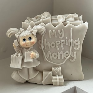 Puede incluir: Una figura blanca de una niña sosteniendo bolsas de compras y un regalo, con una bolsa de compras llena de regalos. El texto "My Shopping Money" está en la bolsa. La figura tiene un collar de perlas y lazos en el pelo.