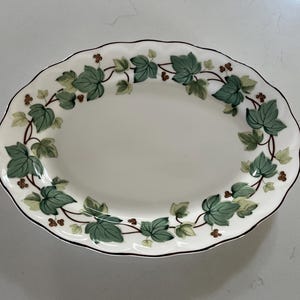 Puede incluir: Plato ovalado para servir con fondo blanco y borde decorativo de hojas de hiedra verdes y detalles marrones. El borde tiene un borde festoneado y una fina línea marrón.