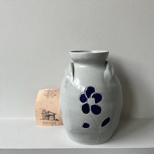 May include: A handmade, off-white ceramic vase with a blue flower design. The vase has a unique shape with handles and a wide opening. A small, aged paper with text is next to the vase.