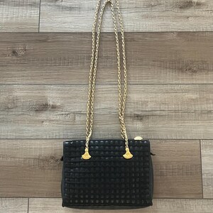 May include: A black rectangular handbag featuring a grid-like textured surface and a gold-tone metal chain strap. The strap is attached to the bag with two gold-tone metal hardware pieces shaped like small fans or tassels at the top corners.