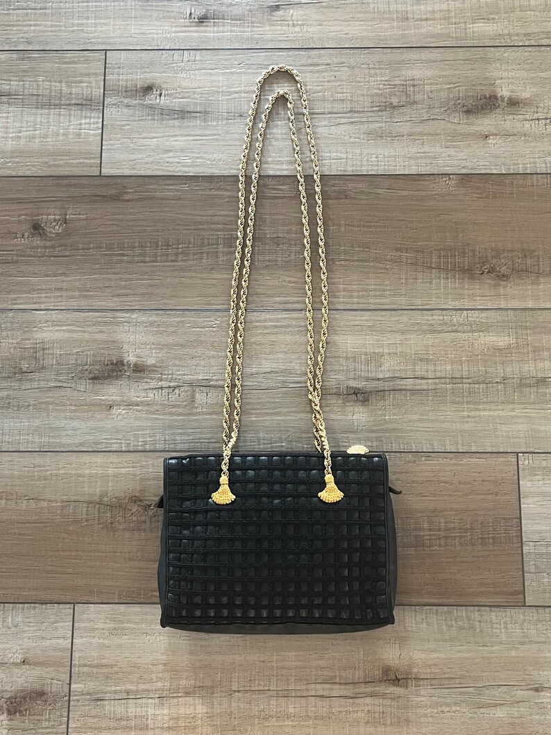 May include: A black rectangular handbag featuring a grid-like textured surface and a gold-tone metal chain strap. The strap is attached to the bag with two gold-tone metal hardware pieces shaped like small fans or tassels at the top corners.
