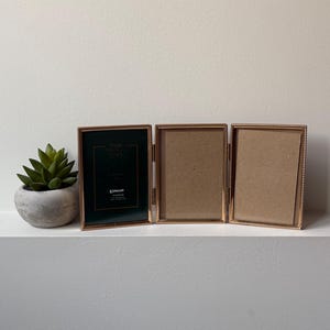 May include: A triptych photo frame with a metallic rose gold finish and beaded edges, featuring three hinged rectangular panels. The left panel contains a black insert with white text, while the two right panels hold plain brown paper inserts.
