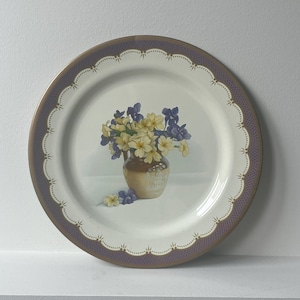 May include: A round decorative plate with a floral design. The plate features a gold rim with a scalloped edge and a purple border. The center of the plate displays a vase with yellow and purple flowers.