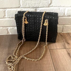 May include: A black rectangular handbag featuring a grid-like textured pattern on the exterior. The bag includes a long, gold-toned twisted rope chain strap that attaches to the top with decorative gold-toned tassels on each side.