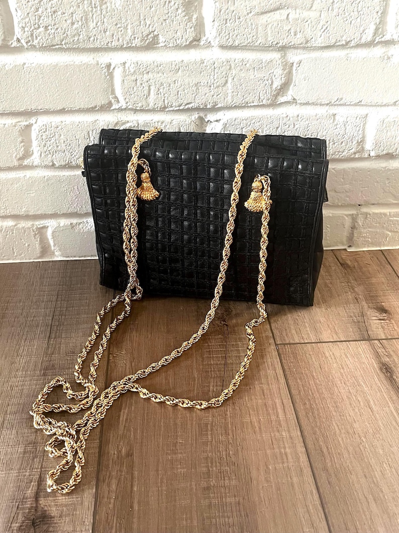 May include: A black rectangular handbag featuring a grid-like textured pattern on the exterior. The bag includes a long, gold-toned twisted rope chain strap that attaches to the top with decorative gold-toned tassels on each side.