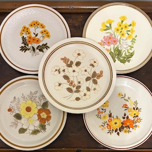 Puede incluir: Un juego de cinco platos redondos de cerámica con diversos motivos florales en tonos amarillo, naranja, rosa y marrón. Cada plato tiene una base de color crema y un borde marrón, con distintas ilustraciones botánicas de flores silvestres.