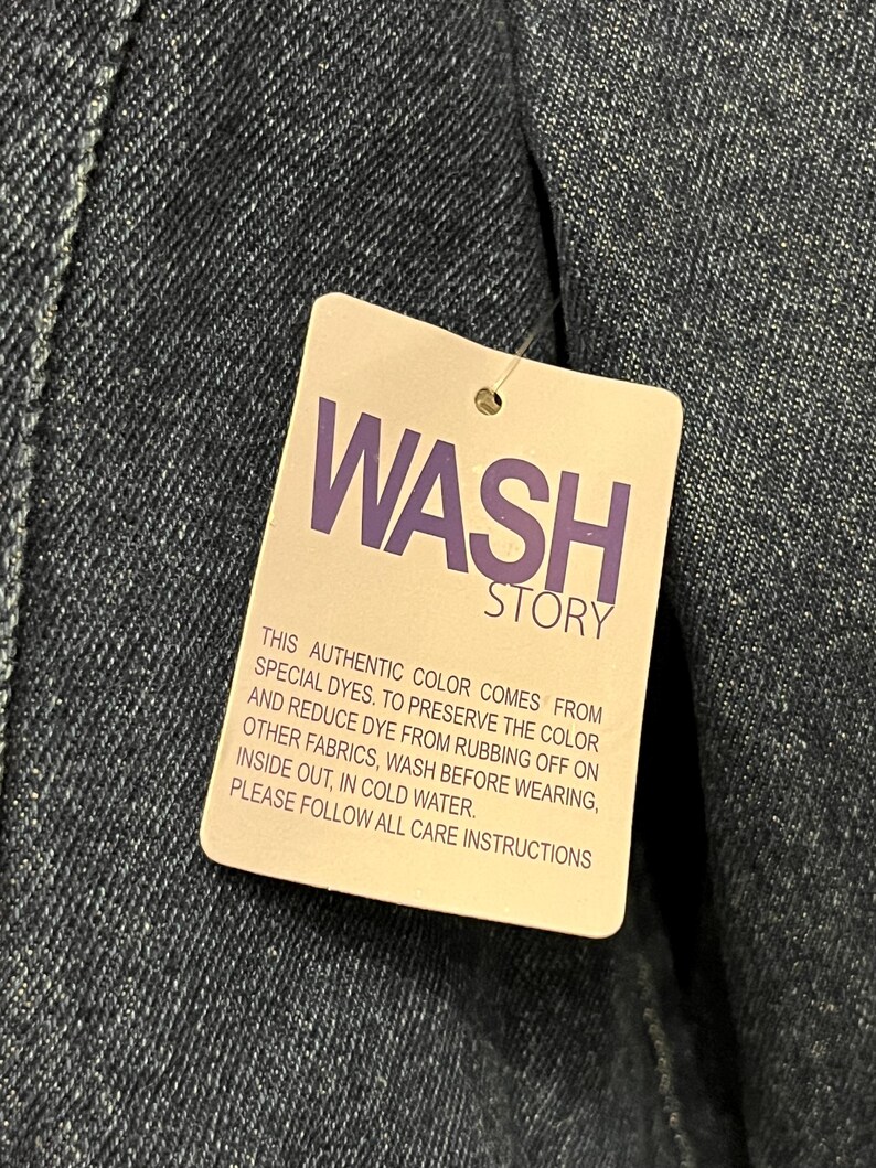 May include: Close-up of a denim garment with a tag that reads "WASH STORY" in purple text. The tag provides care instructions, including washing in cold water and washing inside out.