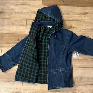 May include: A dark blue denim jacket with a hood and a green and black plaid lining. The jacket has a zipper closure, pockets, and a small tag that says "WASH". The jacket is laid flat on a wooden surface.