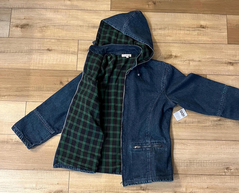 May include: A dark blue denim jacket with a hood and a green and black plaid lining. The jacket has a zipper closure, pockets, and a small tag that says "WASH". The jacket is laid flat on a wooden surface.