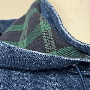 May include: Close-up of a denim jacket collar with a plaid lining in shades of green and navy blue. The jacket's exterior is a medium wash denim, and the collar is reinforced with topstitching. A drawstring is visible.