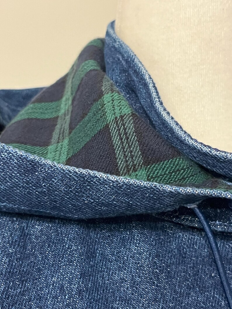 May include: Close-up of a denim jacket collar with a plaid lining in shades of green and navy blue. The jacket's exterior is a medium wash denim, and the collar is reinforced with topstitching. A drawstring is visible.