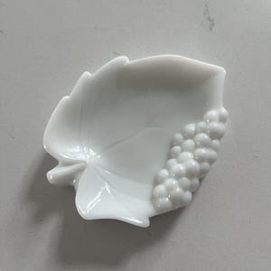 May include: A white, leaf-shaped dish with a cluster of grapes on the right side. The dish is made of a glossy, opaque material. The leaf has detailed veining and a smooth, curved shape. This decorative dish is perfect for serving small snacks or as a decorative accent.