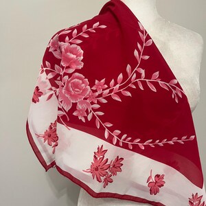 May include: A red and white floral scarf. The scarf features a bold red background with a white border and floral design. The floral pattern includes pink roses and smaller pink flowers with green stems.
