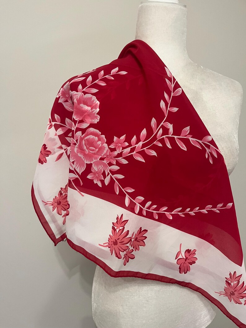May include: A red and white floral scarf. The scarf features a bold red background with a white border and floral design. The floral pattern includes pink roses and smaller pink flowers with green stems.