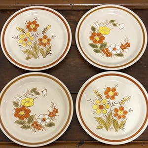Puede incluir: Un juego de cuatro platos redondos de cerámica con base color crema y borde marrón. Dos platos muestran un ramo central de flores naranjas y amarillas con espigas de trigo, mientras que los otros dos presentan un diseño de corona floral en naranja, amarillo y blanco.