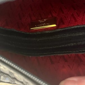 May include: The interior of a black handbag featuring a vibrant red lining with a repeating V-shaped pattern. A rectangular gold-toned metal plaque is centered on the back wall, displaying the text Valentino Garavani in embossed lettering.