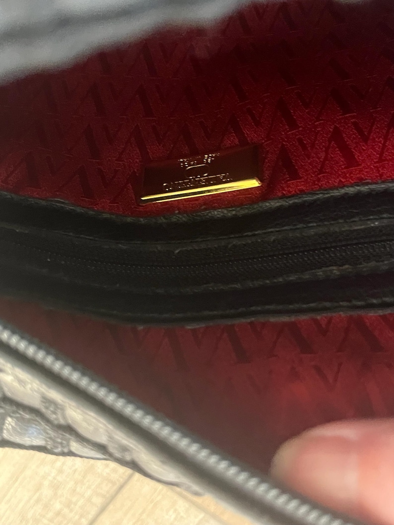 May include: The interior of a black handbag featuring a vibrant red lining with a repeating V-shaped pattern. A rectangular gold-toned metal plaque is centered on the back wall, displaying the text Valentino Garavani in embossed lettering.