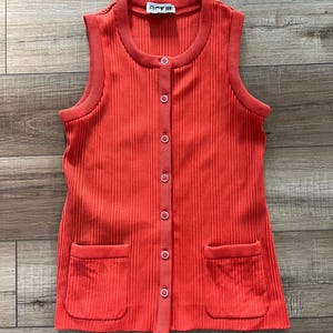 May include: A sleeveless, button-down, ribbed knit vest in a vibrant coral color. The vest features two front pockets and a small tag that reads "ACT III". The garment is displayed on a wooden surface.