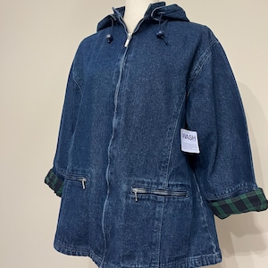 May include: A dark blue denim jacket with a hood and a zipper closure. The jacket has two front pockets with zippers and green and black plaid cuffs. A white tag with the word "WASH" is attached.