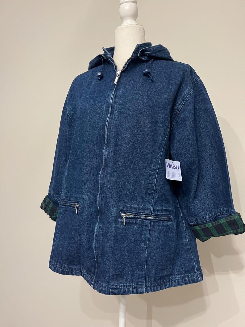 May include: A dark blue denim jacket with a hood and a zipper closure. The jacket has two front pockets with zippers and green and black plaid cuffs. A white tag with the word "WASH" is attached.