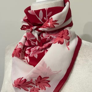 May include: A red and white floral scarf draped over a mannequin head. The scarf features a repeating pattern of pink flowers and leaves against a red and white background. The edges are trimmed with a solid red border.