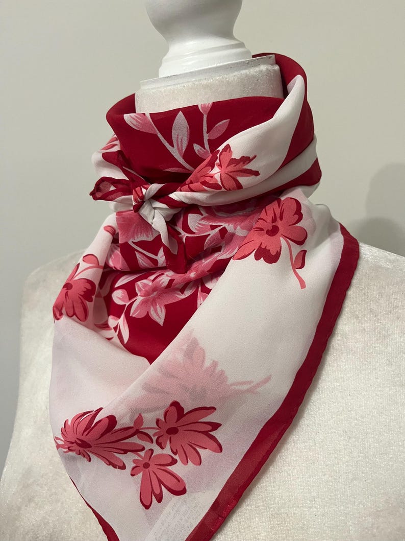 May include: A red and white floral scarf draped over a mannequin head. The scarf features a repeating pattern of pink flowers and leaves against a red and white background. The edges are trimmed with a solid red border.