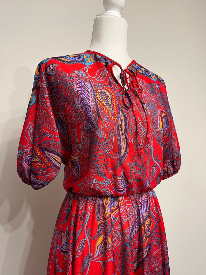 May include: A red, blue, and yellow paisley print dress with a tie at the neck and a gathered waist.