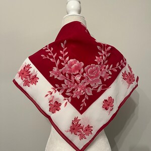 May include: A red and white square scarf with a floral design. The scarf features a red border and a white center with pink flowers and green leaves. The scarf is displayed on a mannequin.