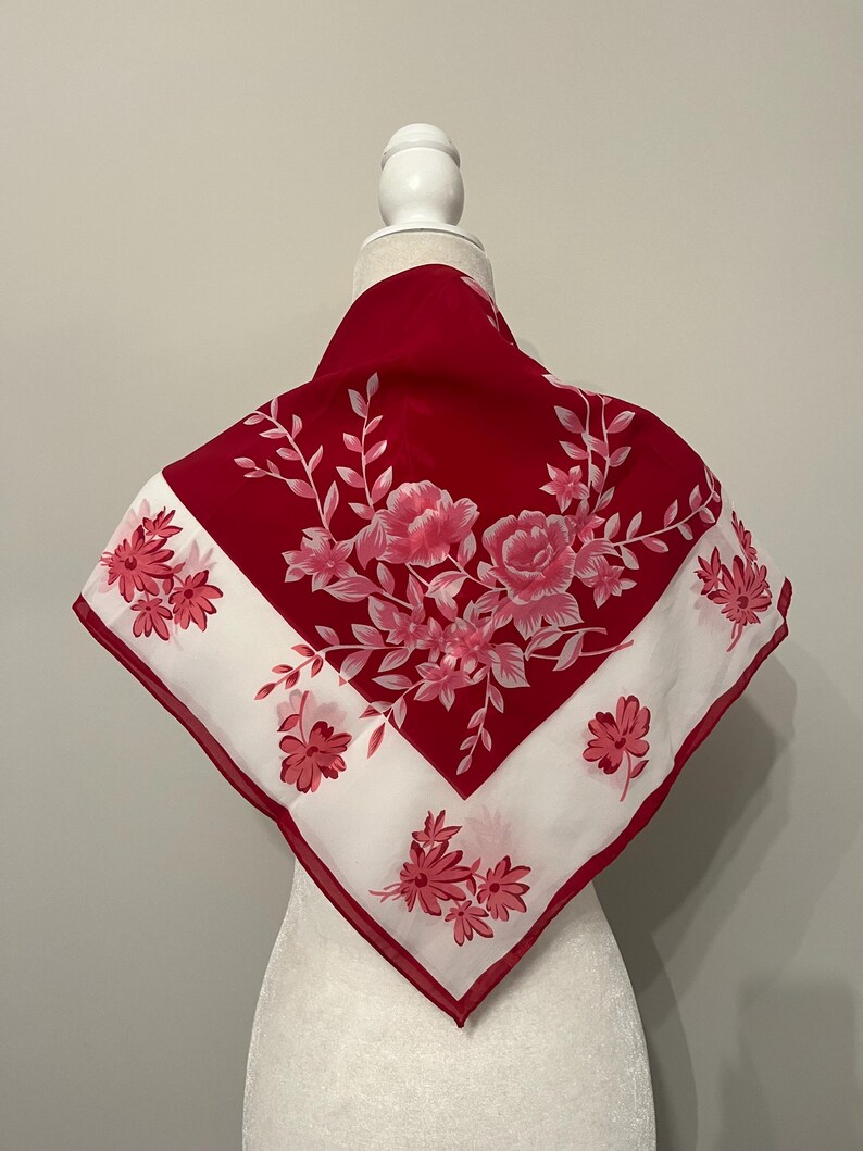May include: A red and white square scarf with a floral design. The scarf features a red border and a white center with pink flowers and green leaves. The scarf is displayed on a mannequin.