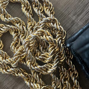 May include: A gold-toned metal chain with a twisted rope-like texture, featuring interlocking links that create a continuous, flexible strand. A portion of a black leather-like material is visible at the edge of the metallic chain links.