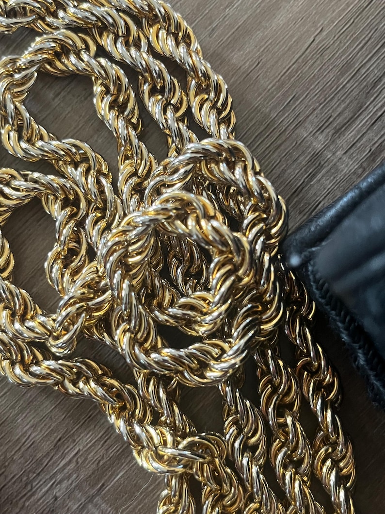 May include: A gold-toned metal chain with a twisted rope-like texture, featuring interlocking links that create a continuous, flexible strand. A portion of a black leather-like material is visible at the edge of the metallic chain links.