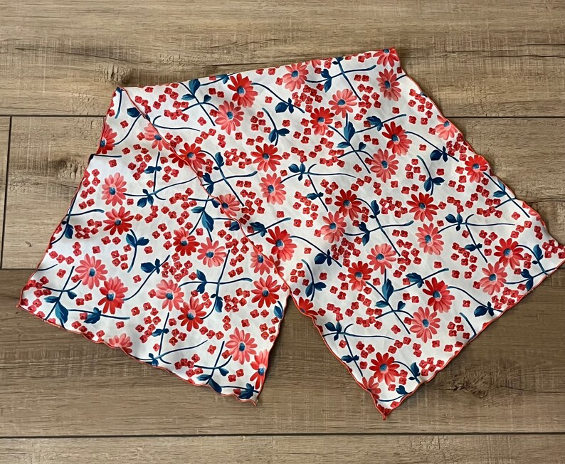 May include: A white fabric scarf with a red and blue floral pattern. The scarf has a scalloped edge and is made of a lightweight material.