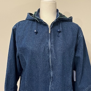 May include: A blue denim jacket with a hood and a zipper closure. The hood lining features a green and blue plaid pattern. The jacket has two front pockets with zippers and blue toggle drawstrings on the hood.