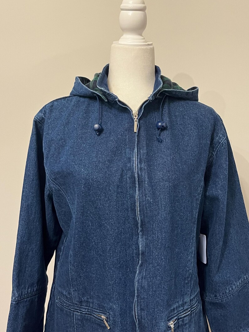 May include: A blue denim jacket with a hood and a zipper closure. The hood lining features a green and blue plaid pattern. The jacket has two front pockets with zippers and blue toggle drawstrings on the hood.