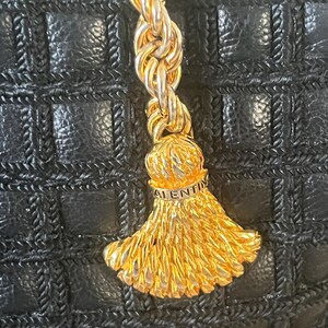 May include: A black handbag with a grid-patterned texture featuring a gold-toned metal rope chain and a decorative tassel charm. The tassel cap is engraved with the text VALENTINO, showcasing a metallic finish against the dark leather surface.