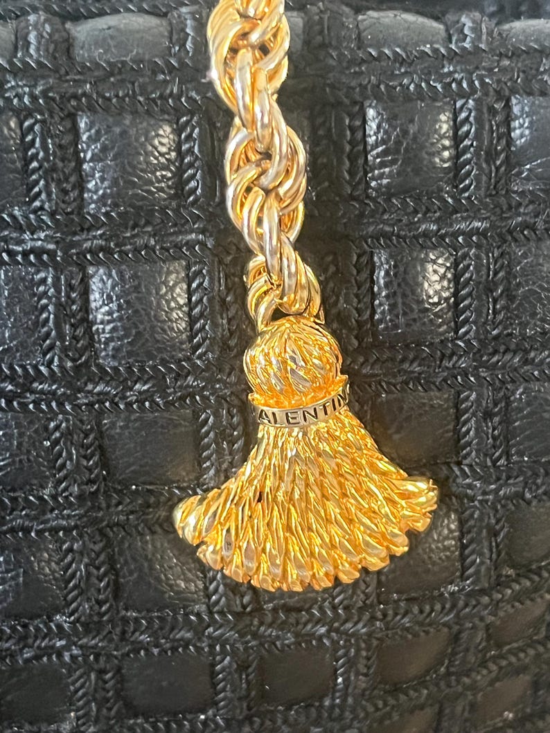May include: A black handbag with a grid-patterned texture featuring a gold-toned metal rope chain and a decorative tassel charm. The tassel cap is engraved with the text VALENTINO, showcasing a metallic finish against the dark leather surface.