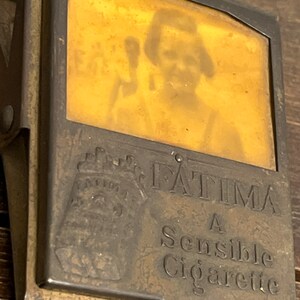 May include: Vintage brass cigarette case with a hinged clip. The case features the name "FATIMA" and the text "A Sensible Cigarette". A faded image of a person is visible through the case's window.