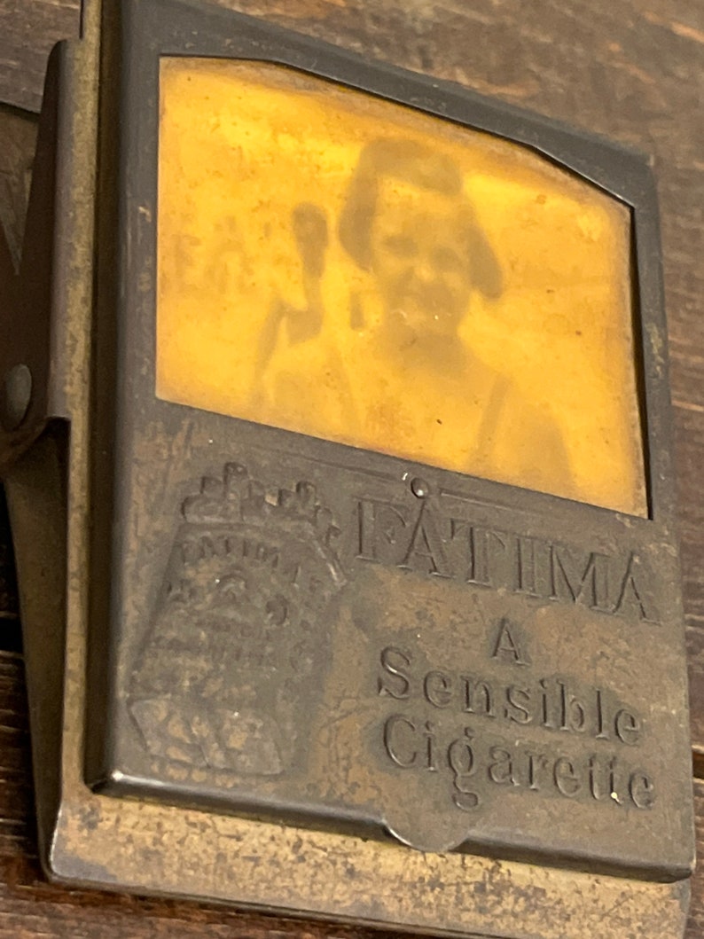 May include: Vintage brass cigarette case with a hinged clip. The case features the name "FATIMA" and the text "A Sensible Cigarette". A faded image of a person is visible through the case's window.